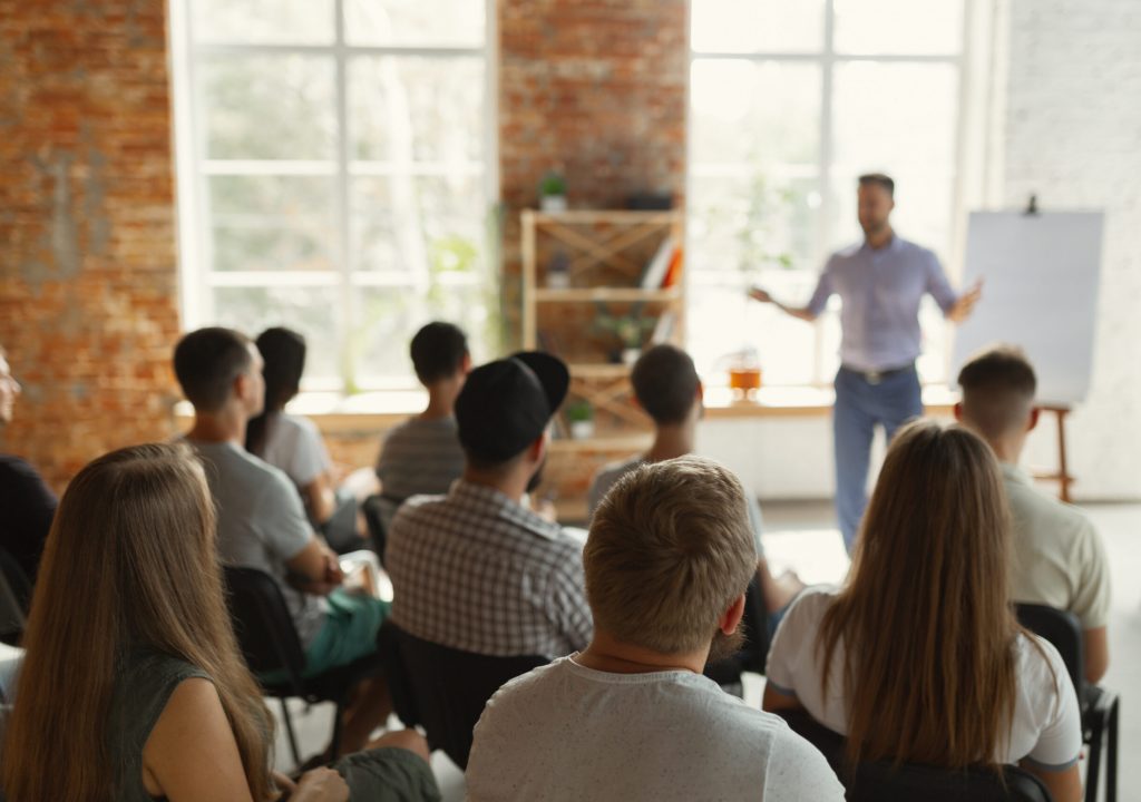 Male speaker giving presentation in hall at university workshop. Audience or conference hall. Rear view of unrecognized participants in audience. Scientific conference event, training. Education concept.